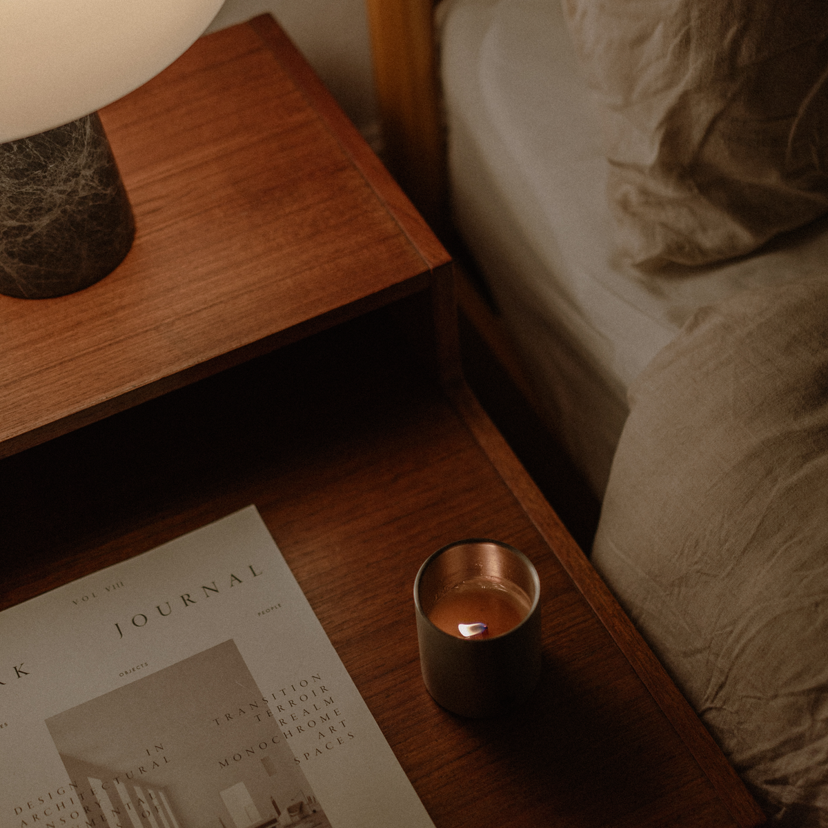 Candle on a bedside table next to a magazine and lamp, with a bed in the background